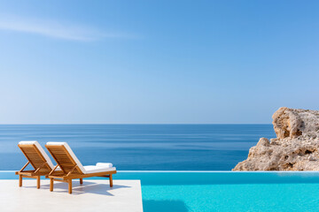 Fototapeta premium Luxurious wooden sun loungers with white cushions by serene infinity pool overlooking calm blue ocean and rocky coastline, perfect for relaxation and tranquility