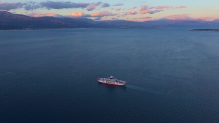 Rio, Greece - 01 December 2024: Aerial view of Rio-Antirrio Bridge at sunset with a ferry on calm sea, Rio, Greece.