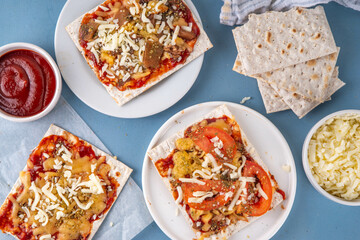 Matzah Pizza on light blue background with ingredients, cheese, tomato sauce. Food ideas and recipe for Jewish Pesach and Passover holidays