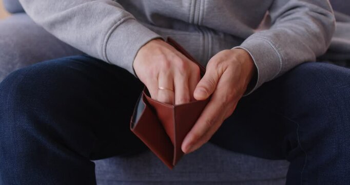 Close up of poor unrecognizable unemployed man wearing casual clothes holding opening empty wallet sitting on sofa at home. Bankrupt, no money, financial crisis and cost control concept. 4k video.