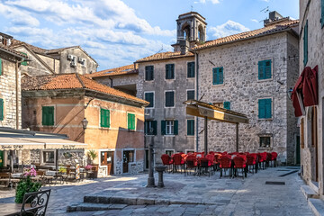 A cozy square in Kotor featuring a charming outdoor cafe with red chairs, surrounded by stone buildings with green shutters. Peaceful vibe of the historic old town. Kotor, Montenegro - June 18, 2021