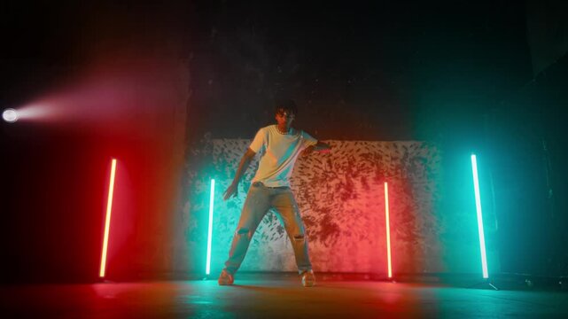 Black b-boy performing active hip-hop dance on stage in dance studio, portrait. African American man enjoying music, moving by rhythm, talented male dancer rehearsing alone in dance studio in night