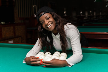 Young black female racking white billiard balls on green pool table, wearing baseball cap and...