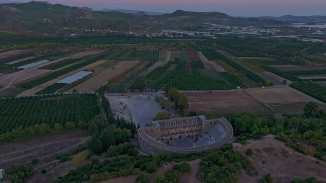 Aerial view of Aspendos Theatre at sunset with surrounding green fields and mountains, Serik, Turkey.