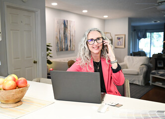 Smiling woman in pink blazer on a phone call at a home office desk.