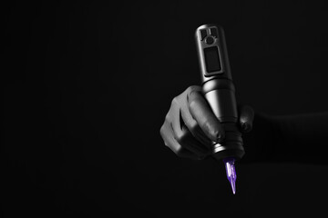 Professional artist in gloves holding tattoo machine on black background, closeup. Space for text