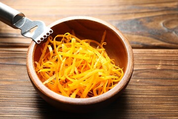 Fresh orange zest in bowl and zester tool on wooden table, closeup