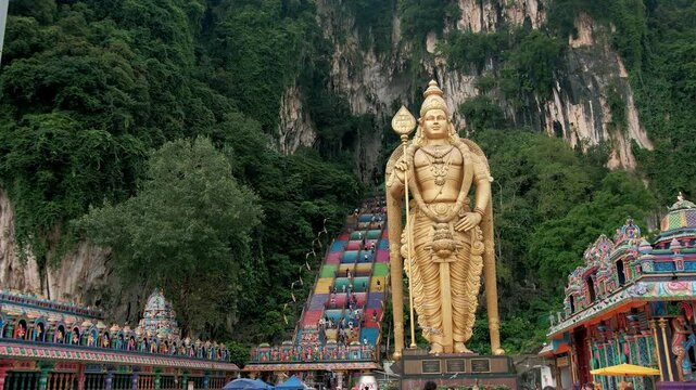 Golden murugan statue greeting visitors near colorful limestone steps leading to sacred hindu temple complex at batu caves, malaysia