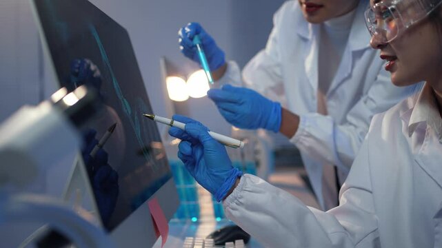 Two scientists in lab coats and gloves examine data on a computer monitor, pointing at the screen with a stylus