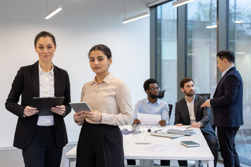 Two female business colleagues are standing in the middle of the office at their workplace, looking seriously and focused at the camera. Meeting and negotiation room.