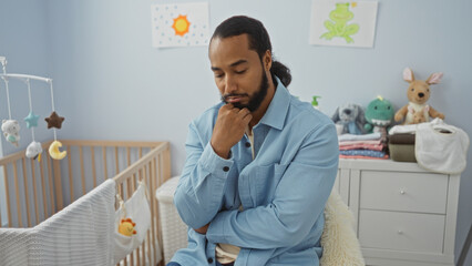 Fototapeta premium Young man with beard seated in a bedroom beside a cradle, appearing thoughtful in the serene, softly lit interior space.