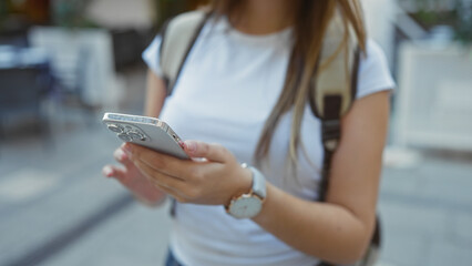 Woman using smartphone while walking on urban street, focused on hands and phone in an outdoor city setting during daytime.