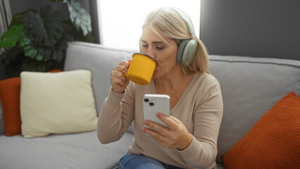 Woman indoors drinking coffee wearing headphones while sitting on a comfortable couch using smartphone in a contemporary living room setting with cozy decor.