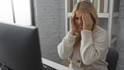 Woman frustrated at workplace, sitting at desk with computer in modern office, covering face with hands, indicates stress or overwork, blonde, female, adult.