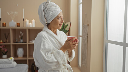 Woman relaxing in a spa room, enjoying a hot drink while wearing a bathrobe with a towel wrapped around her head in a serene and beautiful indoor setting.