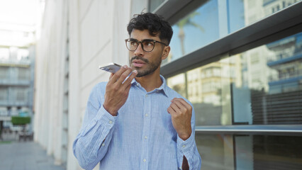 Obraz premium Young man with glasses speaking on smartphone in urban street setting, dressed casually with a beard and surrounded by modern city architecture, exuding confidence and style.