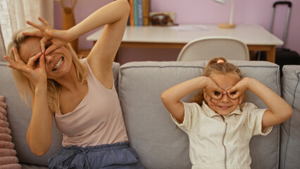 Obraz premium Mother and daughter making silly glasses gesture with their hands in living room