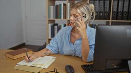 Woman talking on phone while taking notes at desk in office, displaying concentration and...