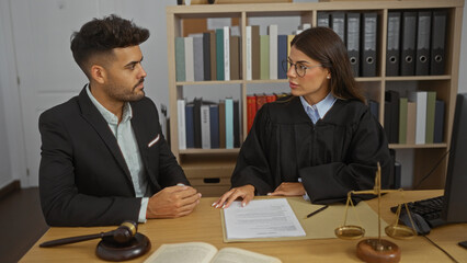 Man and woman working indoors in an office setting, with the woman in judicial robes, discussing documents and law with a judge's gavel and scales in the foreground