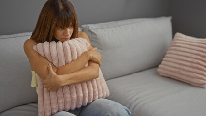 Obraz premium Young hispanic woman with short brunette hair sitting on a couch in a living room hugging a pillow