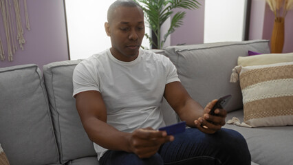 Man sitting on a couch in a cozy living room, holding a credit card and a smartphone, engaged in online shopping, with subtle room decor and bright natural lighting.