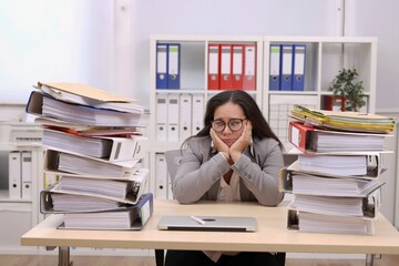female secretary with glasses sits at the desk and is depressed about the piles of files on her desk