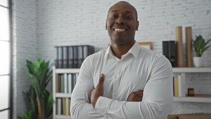 Fototapeta premium Young man with arms crossed in an office room, exuding confidence and professionalism in a modern workplace setting, surrounded by bookshelves and indoor plants.
