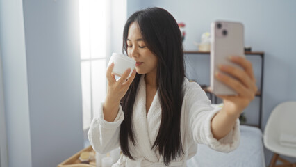 Asian woman taking a selfie and smelling a skincare product in a spa room, emphasizing self-care...
