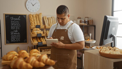Young, hispanic, man working in a bakery shop, taking notes while surrounded by fresh baked bread...