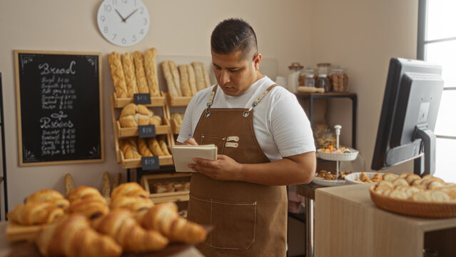 Young, hispanic, man working in a bakery shop, taking notes while surrounded by fresh baked bread and pastries in an indoor setting.