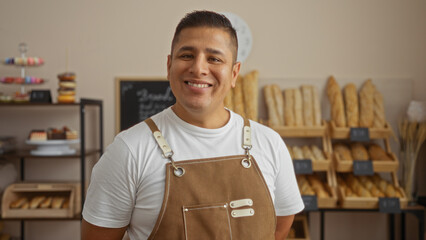 Handsome young hispanic man smiling in a bakery filled with fresh bread and pastries