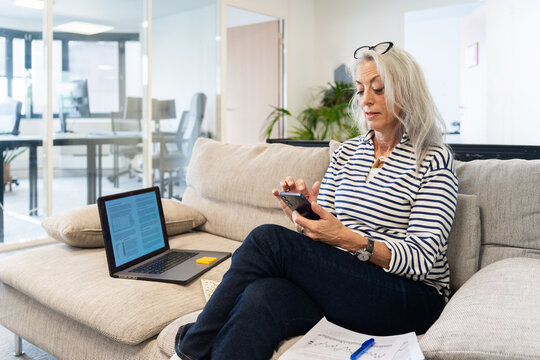 Mature Woman Reviewing Documents and Using Smartphone in Office Lounge