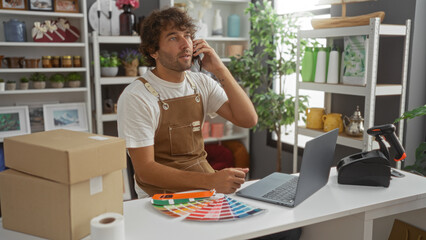 Young man in home decor shop on phone and working on laptop with packages and color samples on table