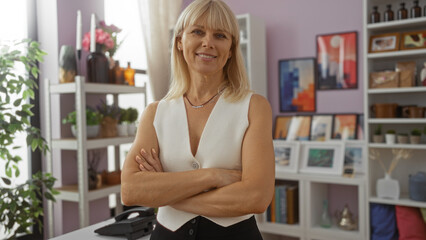 Beautiful caucasian mature woman standing in a home decor store with folded arms and smiling confidently amid various decorative items, art pieces, and shelves.