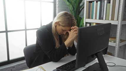 Woman stressed at computer in office with blonde hair sitting indoors under natural light mourning...