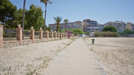 Obraz premium Outdoor scene with blurred pathway leading to buildings surrounded by trees, fenced area, palm trees, and sandy ground, under a clear blue sky
