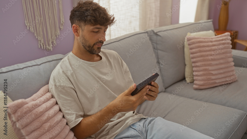 Wall mural Young man sitting on a couch at home looking at a picture frame with pillows around him in a cozy, indoor living room