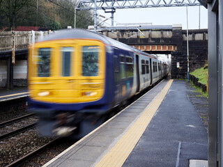 Blurred train speeds through a railway station under an overpass on a rainy day.