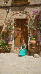 Young woman sitting in front of rustic house in valldemossa, mallorca, surrounded by blooming flowers in outdoor setting under bright sunlight