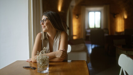 Young hispanic woman sitting at a table in a cozy indoor restaurant, gazing out of a window with a calm expression, surrounded by warm lighting and a homey atmosphere