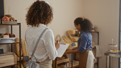 Two latin women working in a bakery interior, one taking notes with clipboard while the other prepares bread
