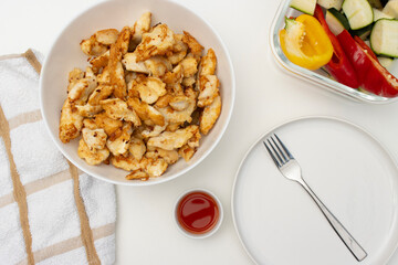 On a white table there is a bowl with fried chicken, a baking tray with vegetables and a plate with a fork. Homemade dinner on a white background. Copy space