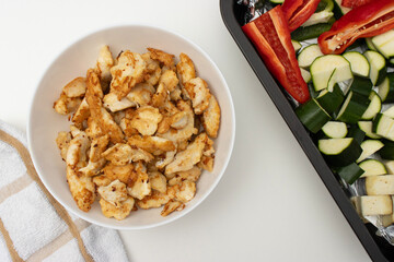 On a white table there is a bowl with fried chicken, a baking tray with vegetables and a plate with a fork. Homemade dinner on a white background. Copy space