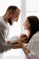 Fototapeta premium Portrait of a young family with infant in his arms photographed on a light window background.