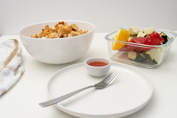 On a white table there is a bowl with fried chicken, a baking tray with vegetables and a plate with a fork. Homemade dinner on a white background. Copy space