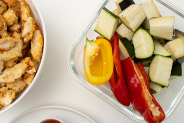 On a white table there is a bowl with fried chicken, a baking tray with vegetables and a plate with a fork. Homemade dinner on a white background. Copy space