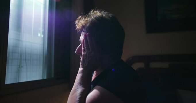 Elderly woman sitting near window resting hands on face feeling anxiety and quiet despair introspective moment of angst serene indoor atmosphere of solitude