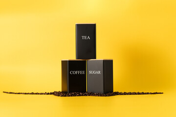 Black containers for tea, coffee, and sugar on a yellow background, with some wood in the caps 
