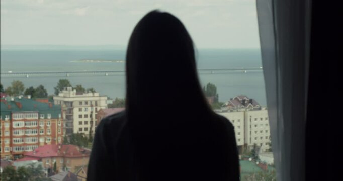 Woman opening curtains and looking at cityscape by the sea