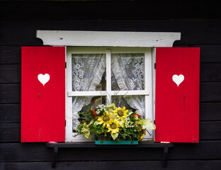 A charming window with red shutters featuring white heart shapes, adorned with lace curtains and a colorful flower box filled with sunflowers and greenery.
